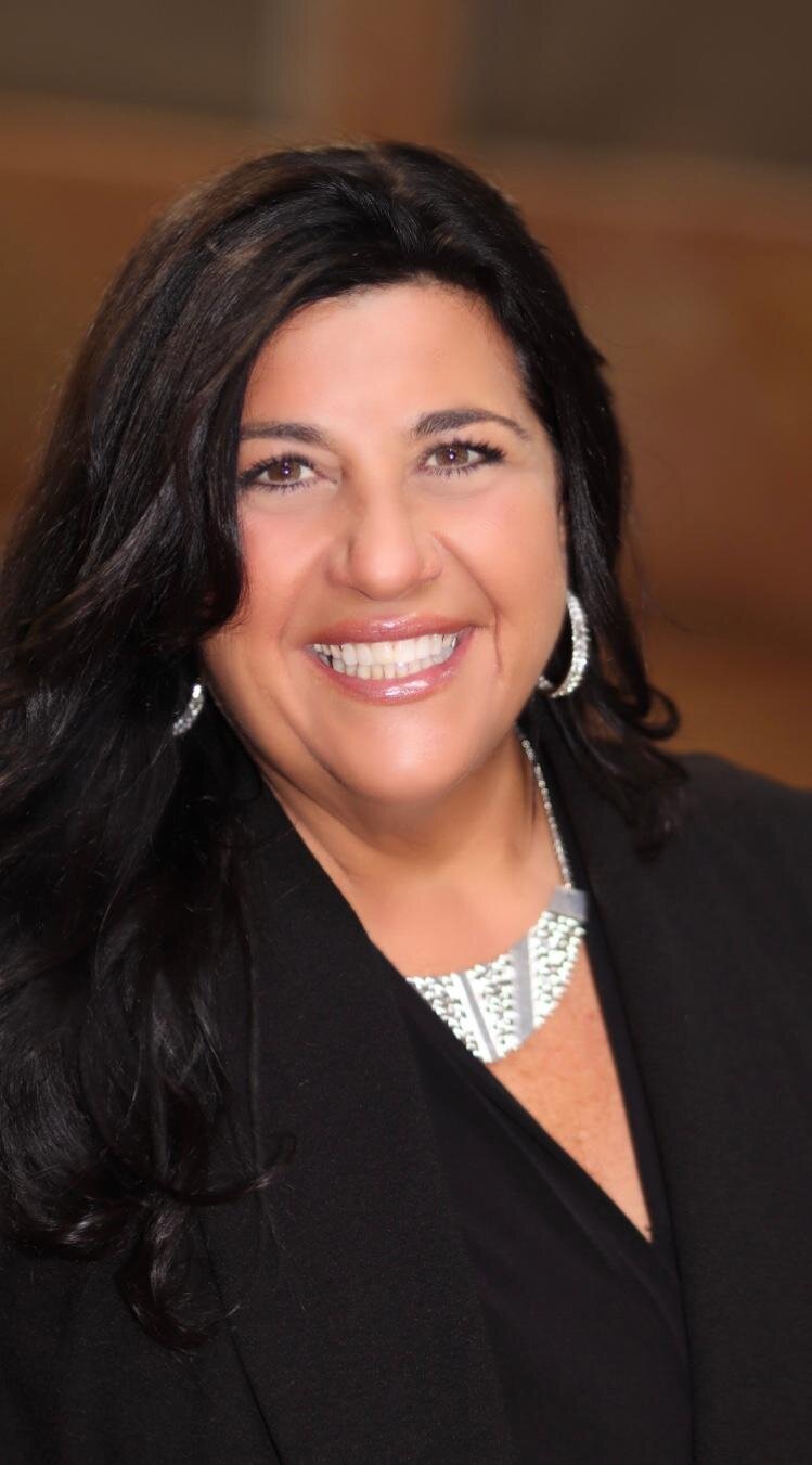Close-up of a woman with long dark hair, wearing a black top and jewelry, smiling at the camera.