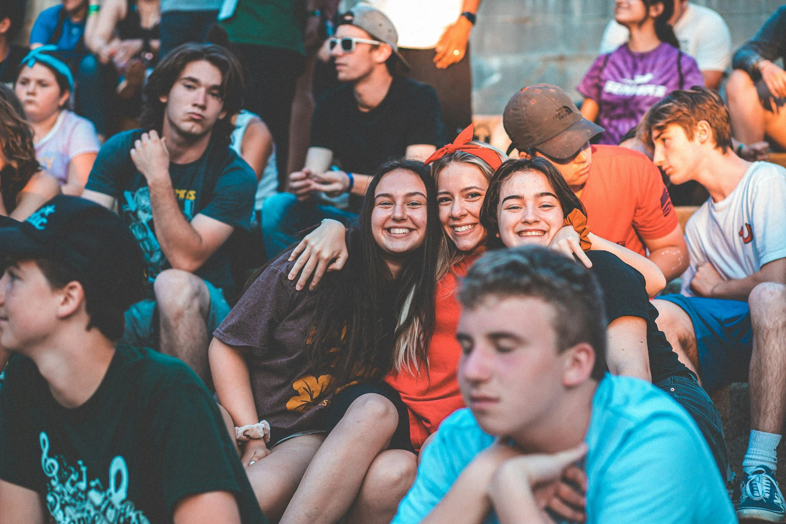 A group of young people sitting outdoors, with three girls in the center smiling and hugging each other.