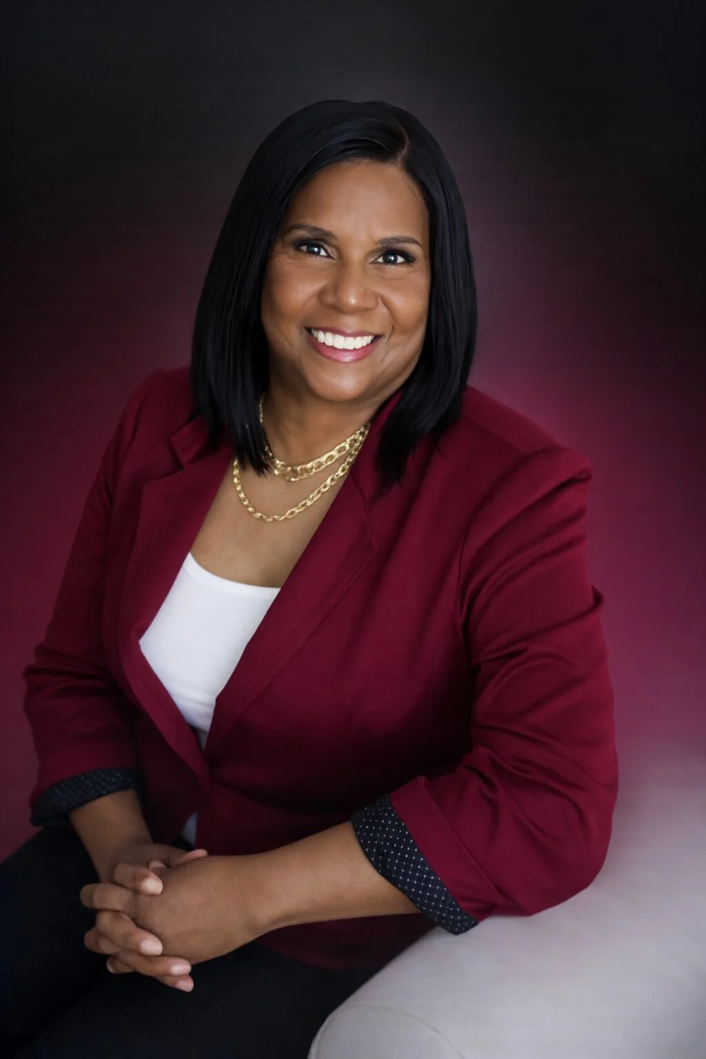 A professional woman with dark shoulder-length hair, wearing a maroon blazer, white top, and gold jewelry, smiling at the camera against a dark background.