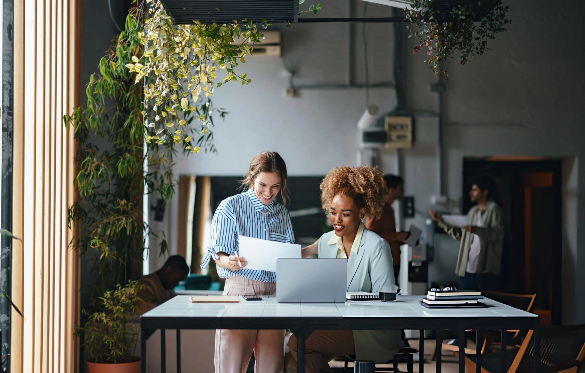 Two women working together at a desk in a modern, green office space. One woman is standing and pointing at papers, the other is seated, looking at a laptop. Other people are working in the background.