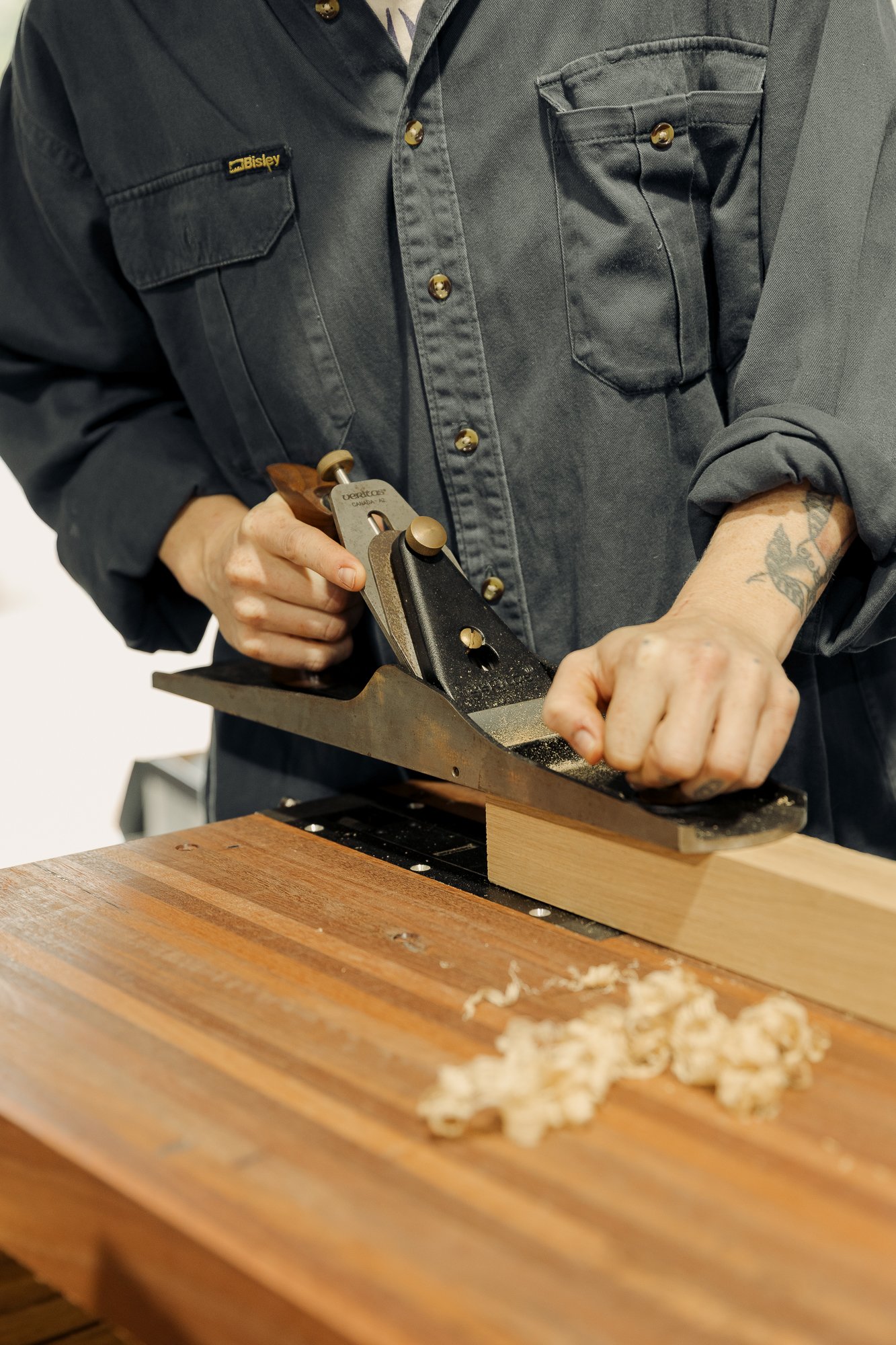 Person wearing a dark gray shirt using a hand plane to smooth a piece of wood on a workbench.