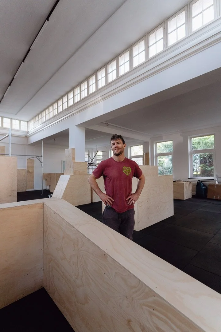 Simon Pouly steht stolz in der frisch ausgebauten Parkourhalle mit schwarzem Gummiboden und aus Holz gefertigten Parkourhindernissen. Die schöne Halle hat viele Fenster.