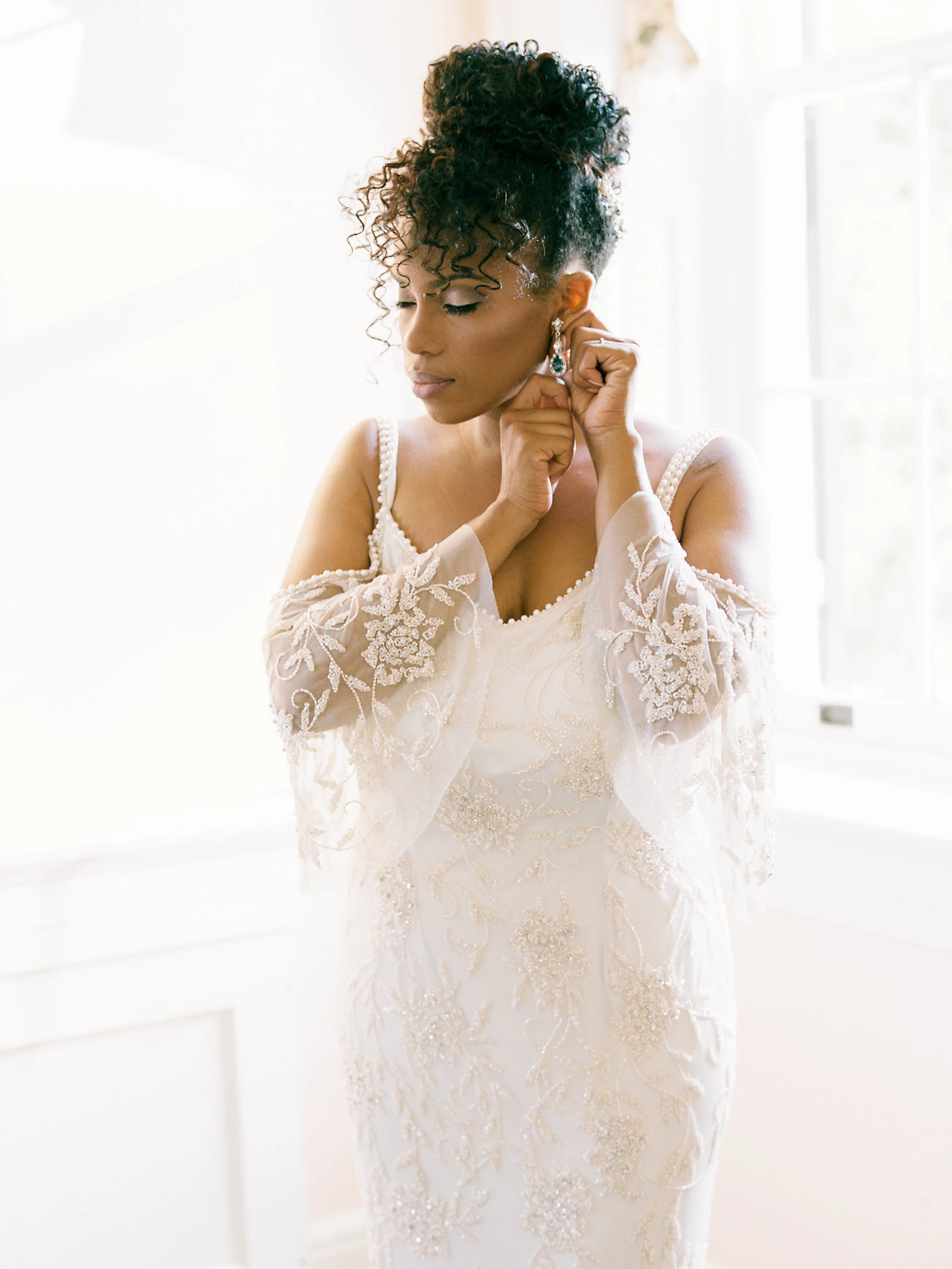Bride in a lace gown putting in her earrings, backlit by bright window light