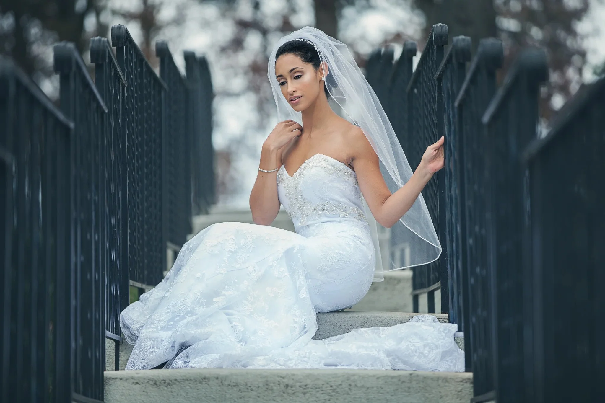 Bride in a strapless wedding gown and veil seated by an iron gate, bridal makeup by Keena Queen