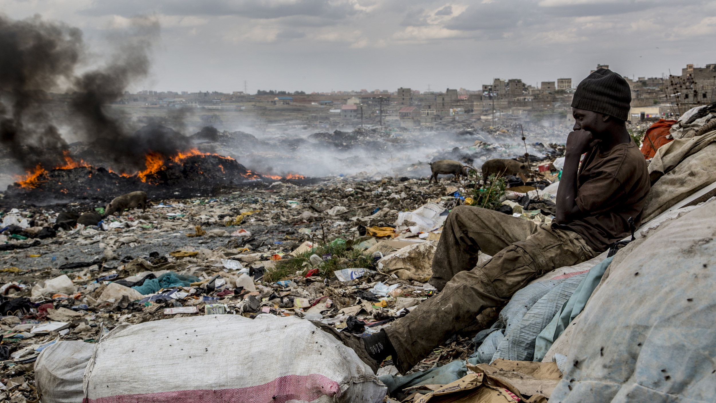  Benson Mumo Muli, 21, watches over a fellow colleague's cache as a mound of trash burns in the background. Benson has been sorting and selling plastics at the site for the past two years. He also fashions what appeared to be reams of plastic (think 