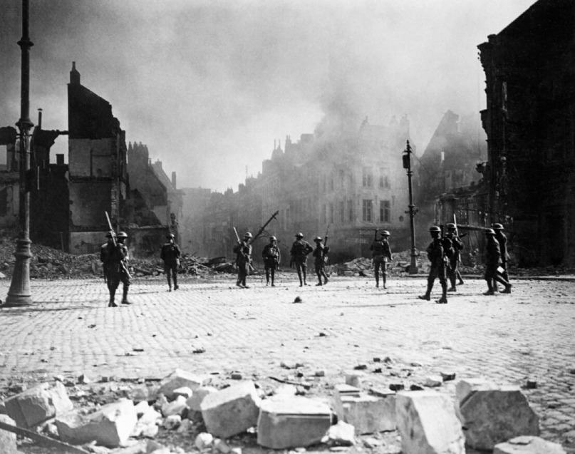 A group of Canadian soldiers strung out in the streets of Cambrai against its still burning buildings. Photo by William Rider-Rider (Imperial War Museum, CO3373)
