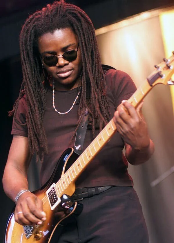 Tracy Chapman is wearing sunglasses and looking down at her guitar as she plays at a concert.