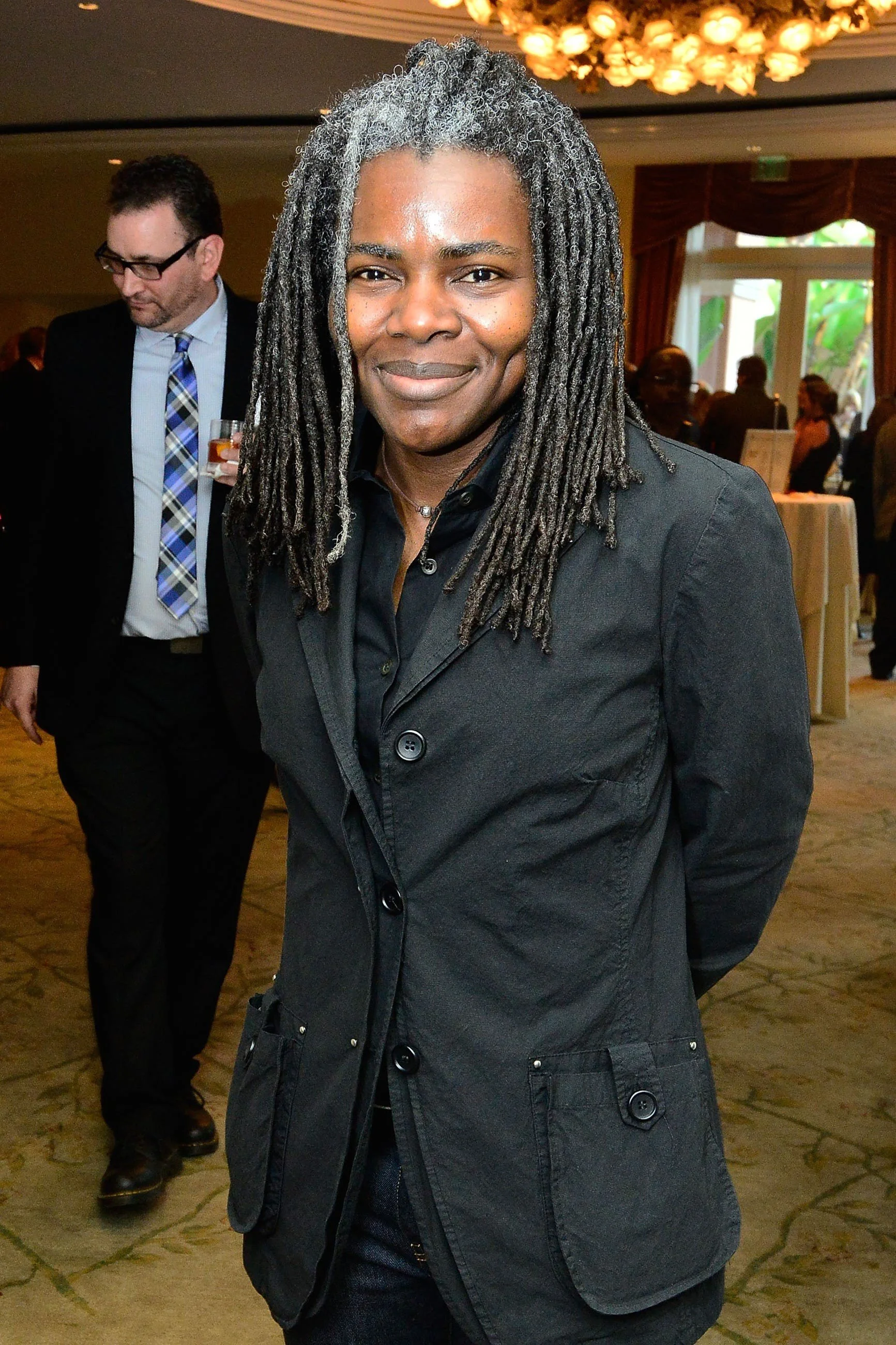 Tracy Chapman is wearing a suit and smiling at the camera with her hands behind her back.