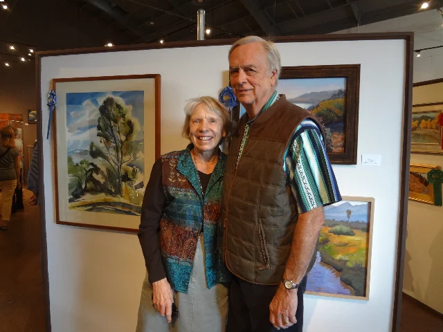 Guest Juror Patti Jacquemain and husband Dave against wall of award winning paintings on display thru Oct. 4th