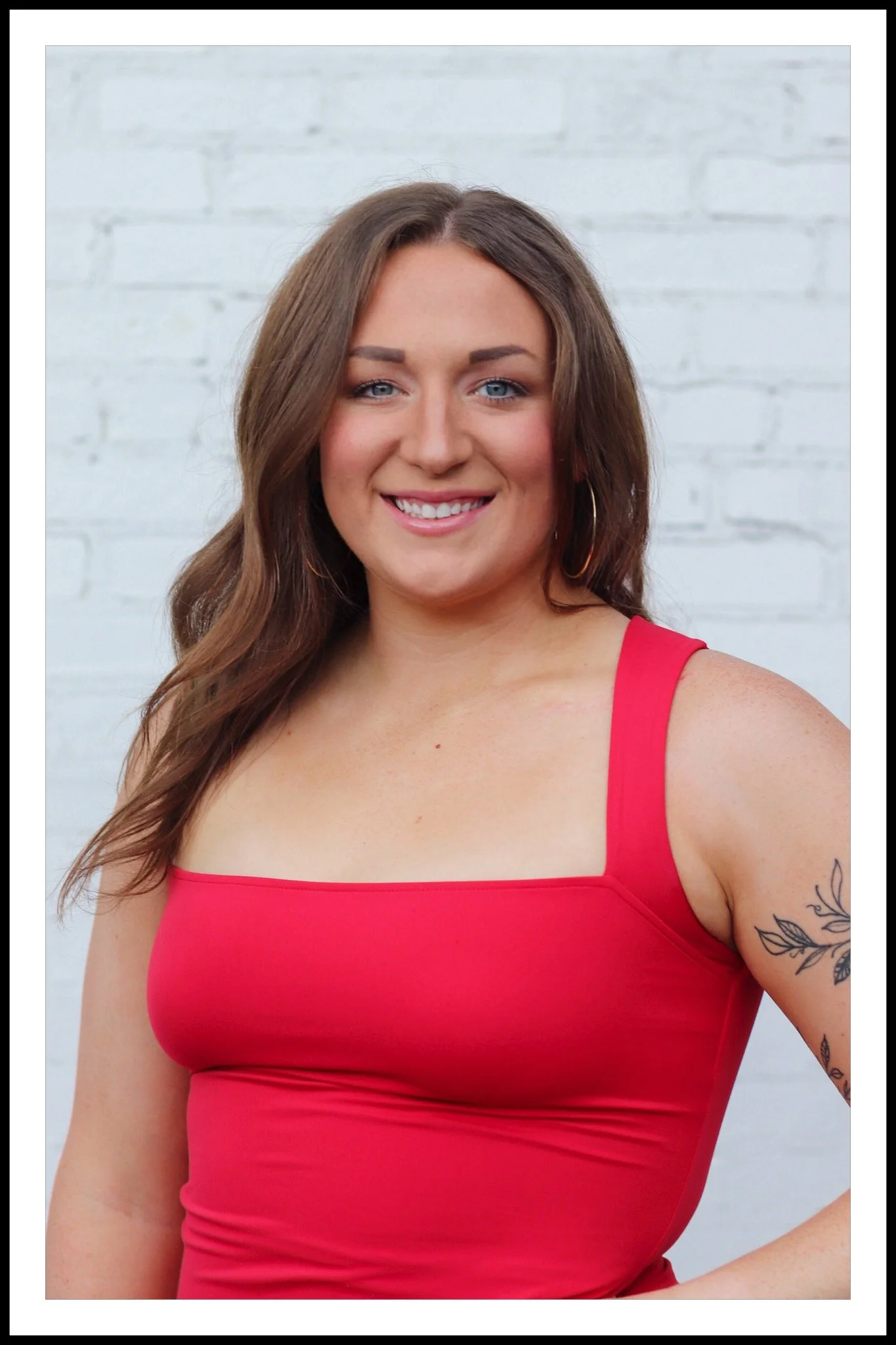 A smiling woman with long brown hair and blue eyes, wearing a red sleeveless top, standing in front of a white brick wall.