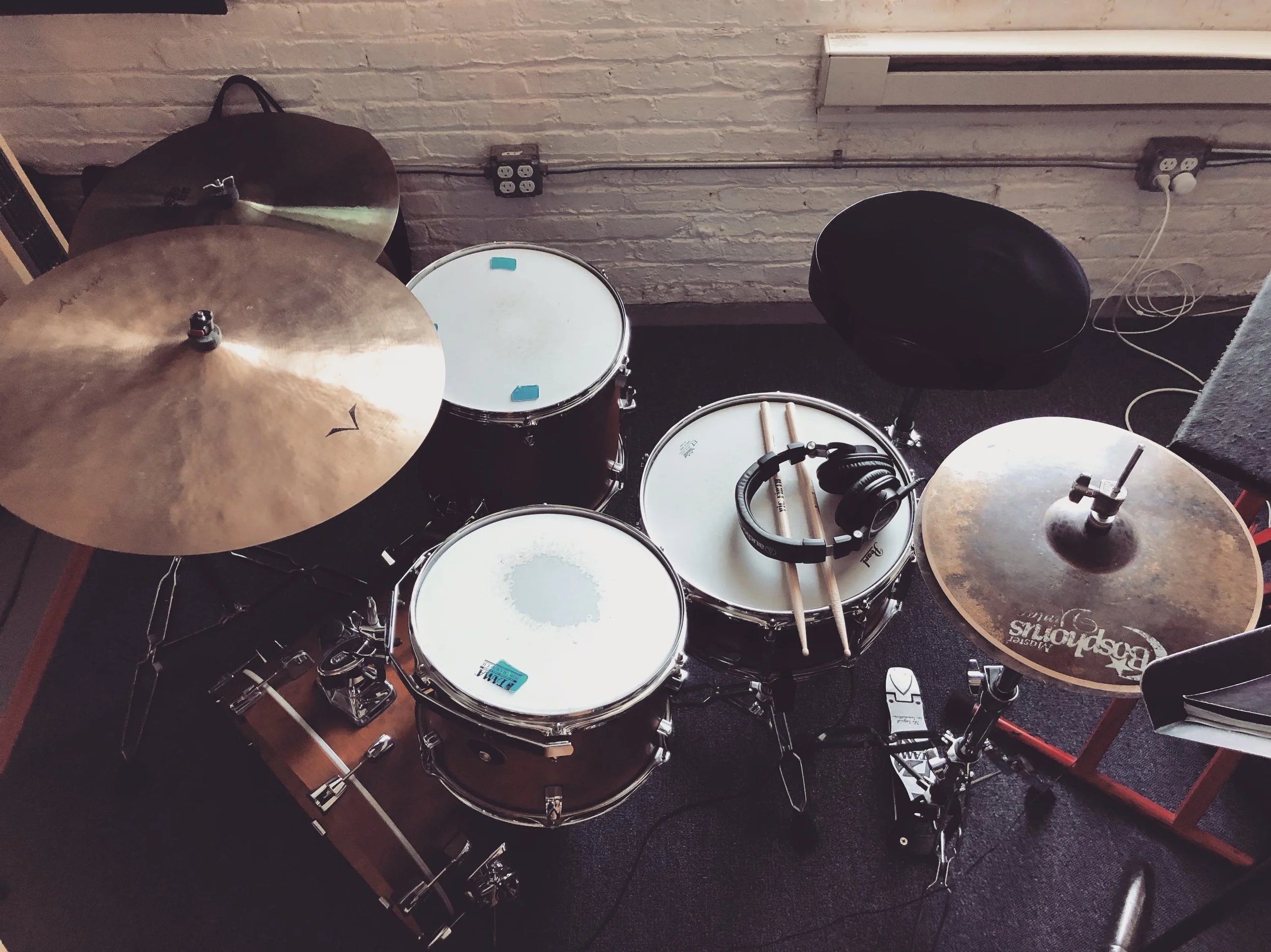 A drum set with cymbals, snare drum, tom-toms, and a bass drum, placed on a black carpet in front of a white brick wall. There are headphones resting on the drums and some wires and a black drum throne nearby.
