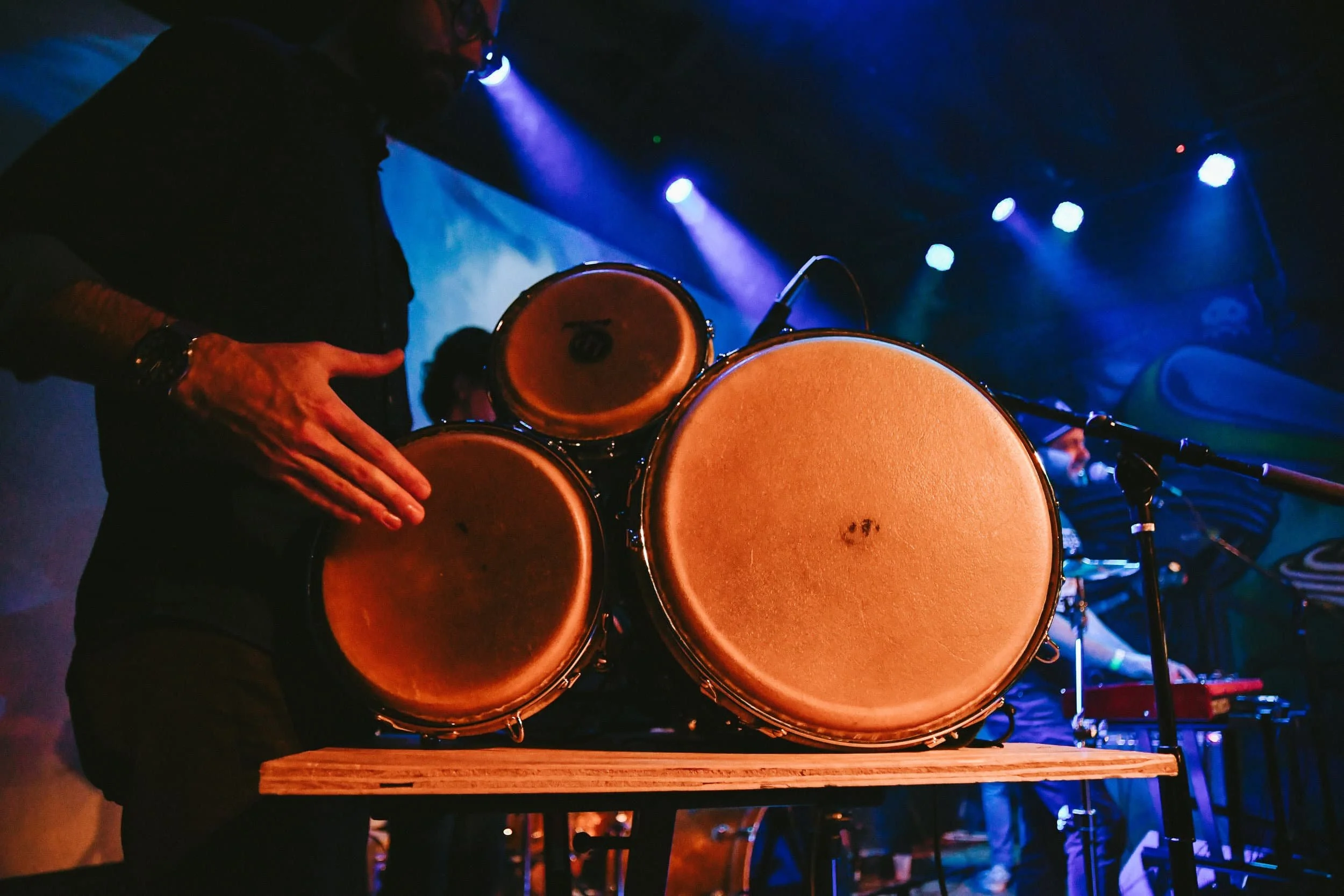 A person playing bata drums on a wooden surface, with colorful stage lighting and a dark background.