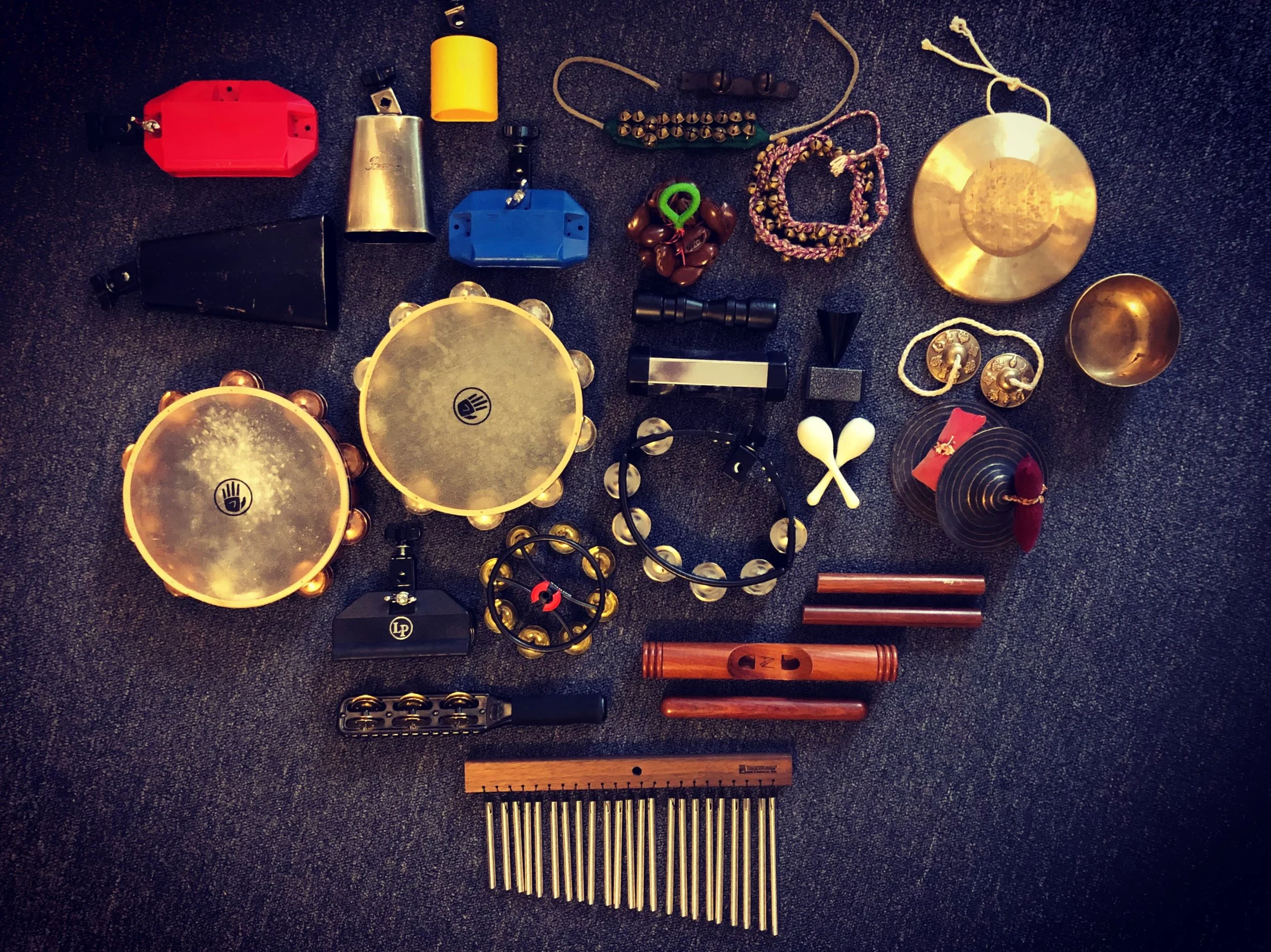 Collection of musical instruments and accessories on a dark carpeted surface, including tambourines, claves, windchimes, a shaker, and cowbells.