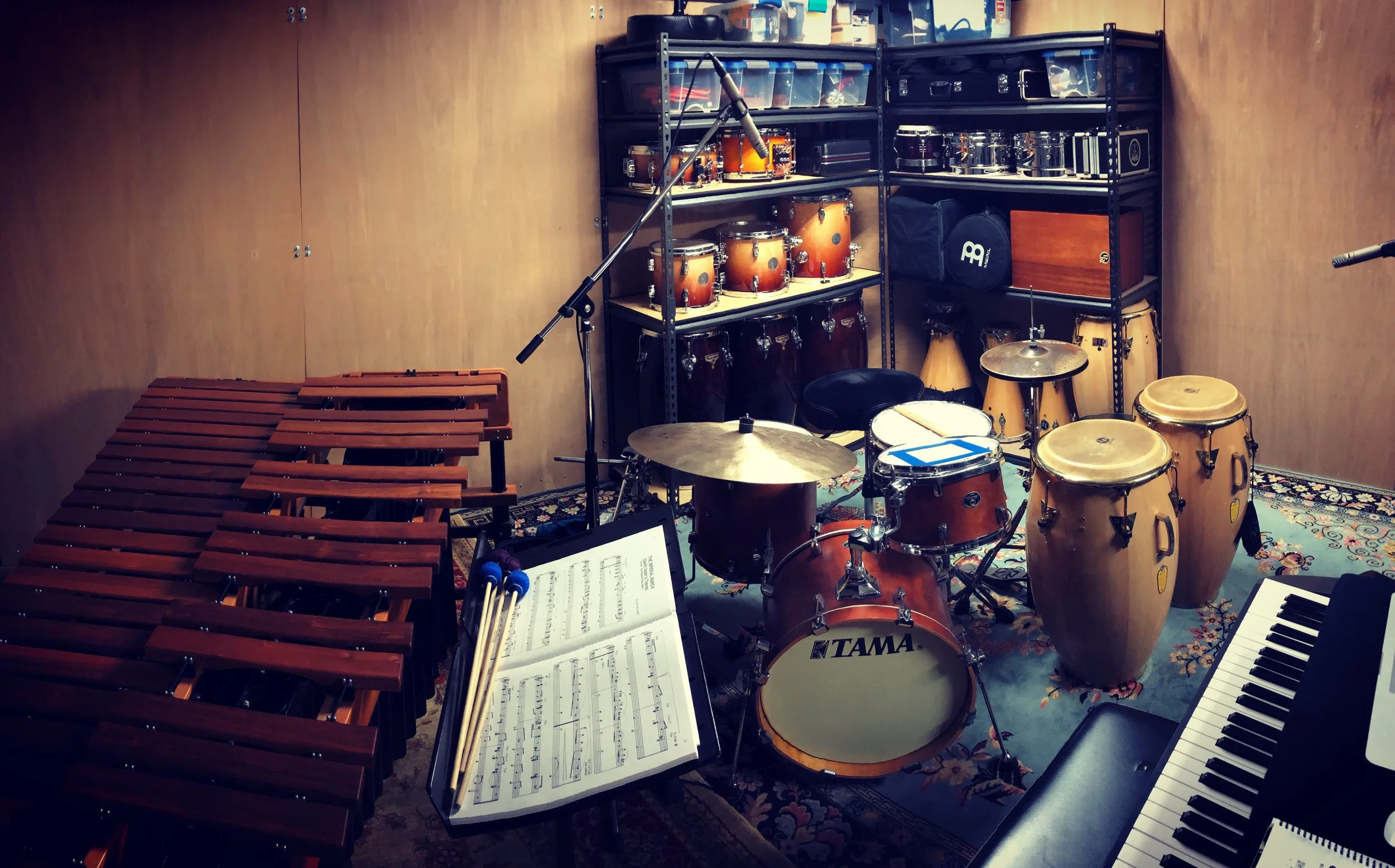 A music rehearsal room with various percussion instruments, including a marimba, drums, congas, and a keyboard, along with sheet music and microphones.