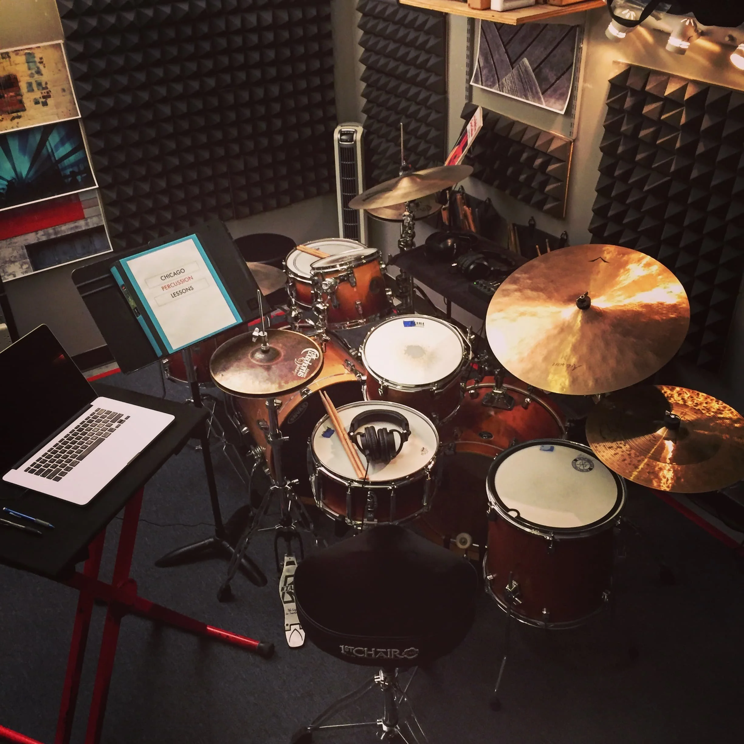 Music practice room with acoustic foam panels, a drum set, a music stand with a playlist titled 'Chicago Percussion Lessons,' a laptop, and a chair, all on a dark carpeted floor.