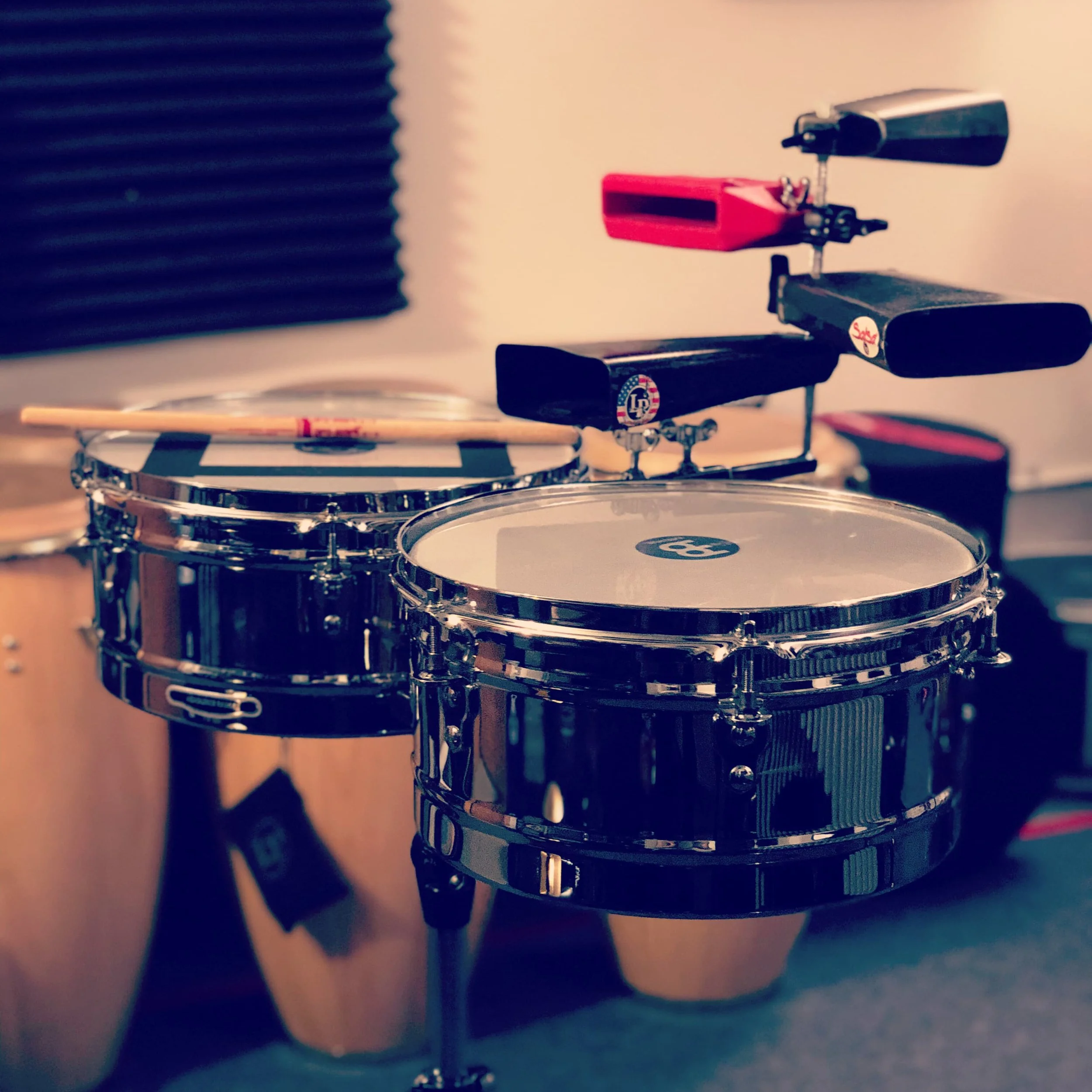Close-up of timbales, with metal rims, cowbells, and drumsticks resting on it, and soundproof foam panel in the background.