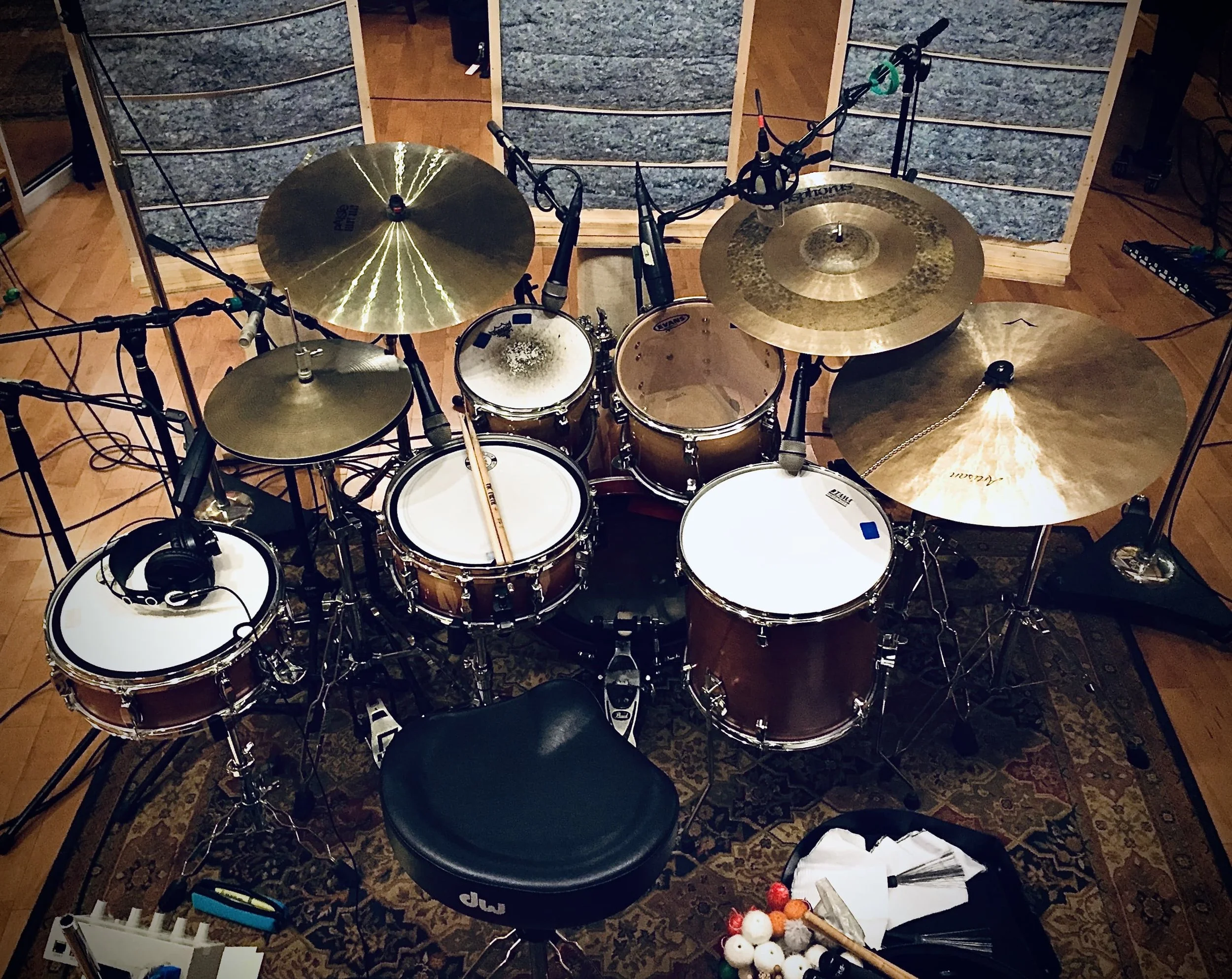 A top-down view of a drum set and cymbals on a wooden floor with various microphones and cables, a black stool, and some drumsticks and percussion accessories nearby.