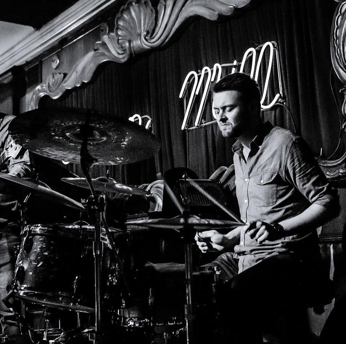 A man playing drums in a dimly lit venue with a neon sign in the background.