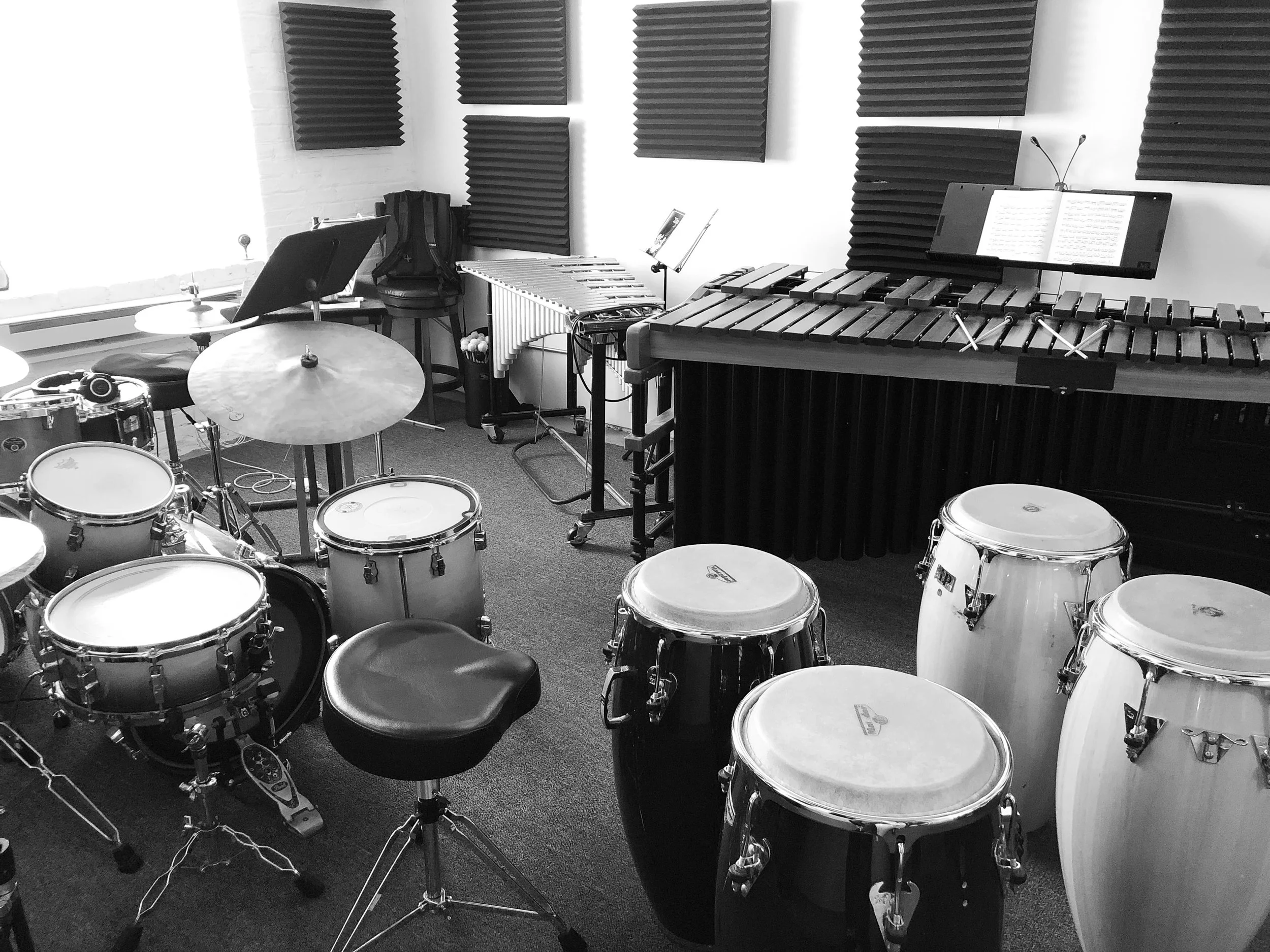 A music rehearsal room with drums, a marimba, congas, a music stand with sheet music, and acoustic foam panels on the walls.