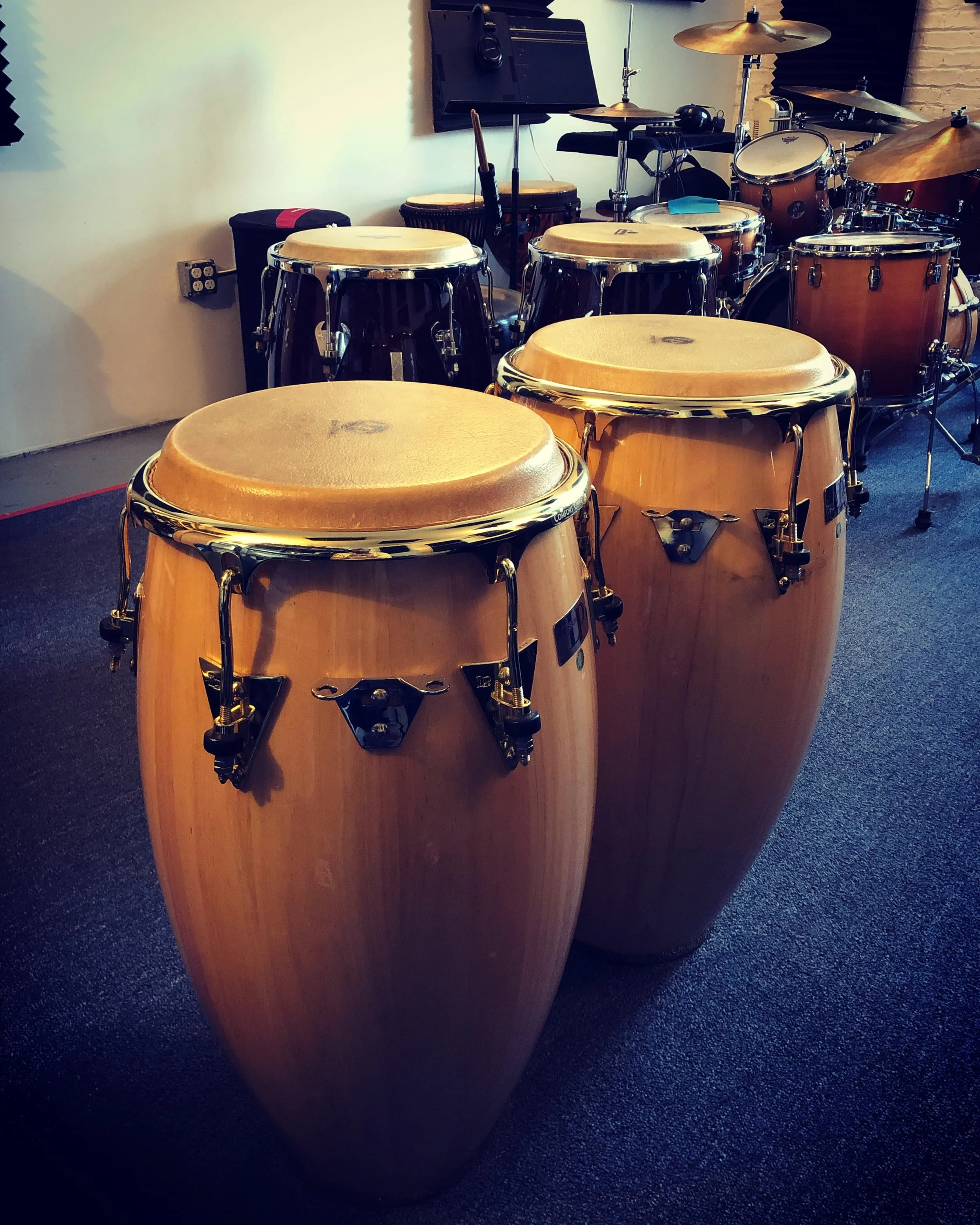 Close-up of a set of wooden conga drums with metal hardware, positioned on a dark carpeted floor in a music studio or practice space.