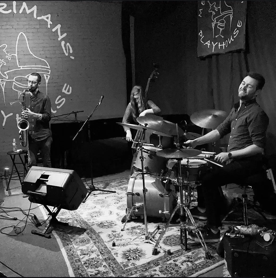 A three-member jazz band performing live on a stage, with one musician playing the saxophone, another on the drums, and a third on the upright bass, in a dimly lit venue.