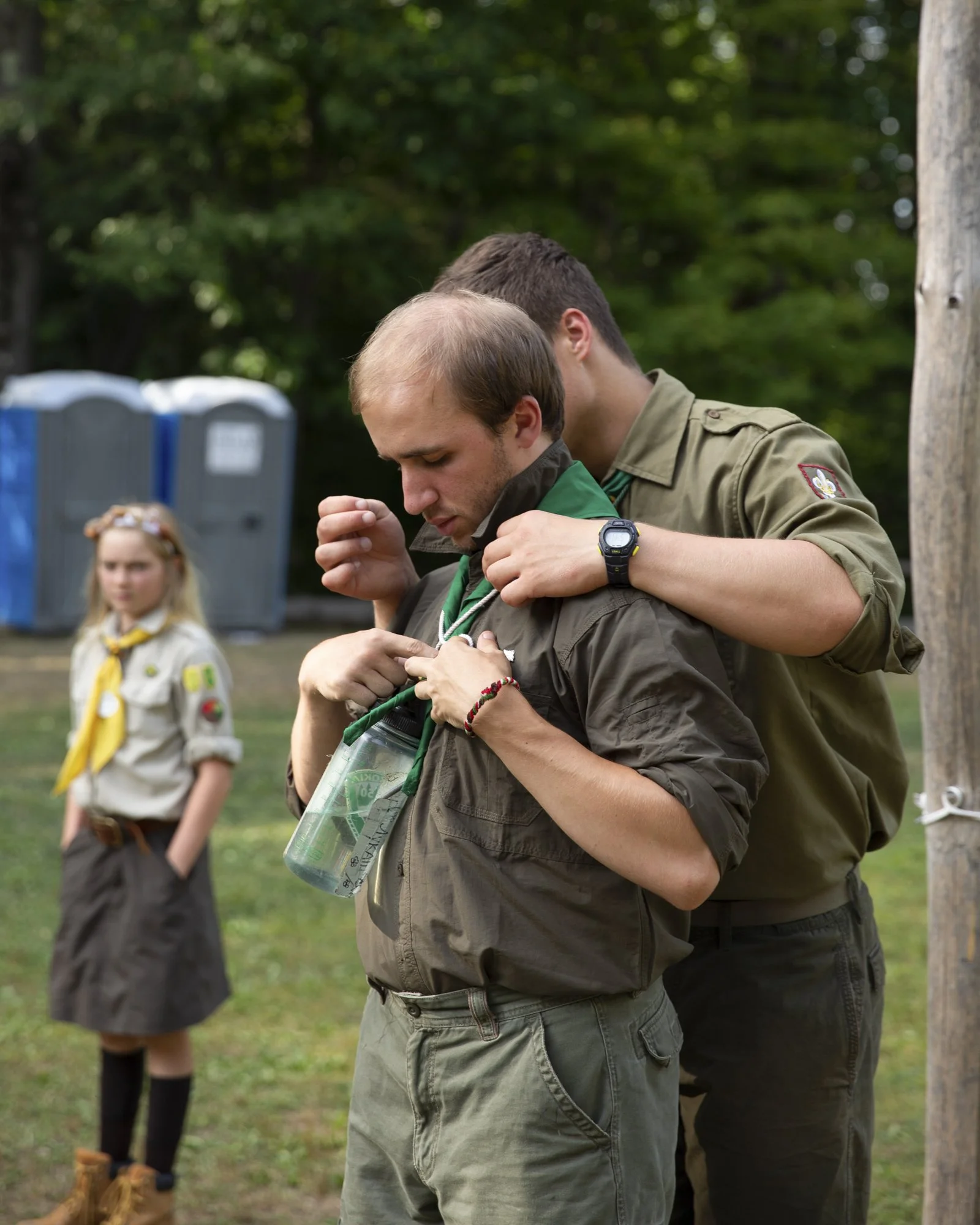 A young adult man, possibly in his 20s or early 30s, is being helped by another young man to adjust his neckerchief in a scout uniform. In the background, a young girl in a scout uniform with a yellow neckerchief watches; all appear to be in an outdo