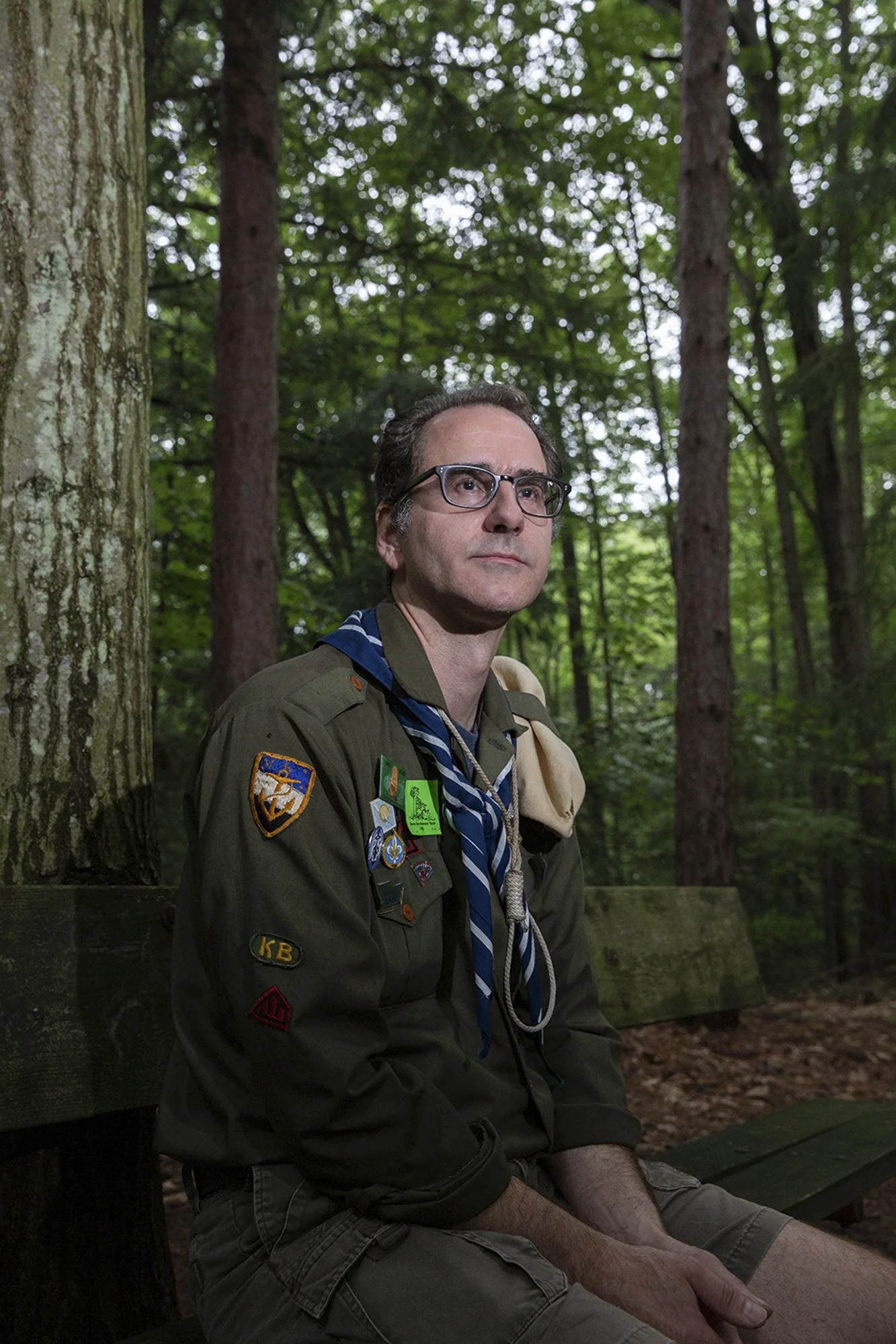 An adult male Caucasian scout leader wearing a uniform with badges looks thoughtfully into the distance in a forest setting.