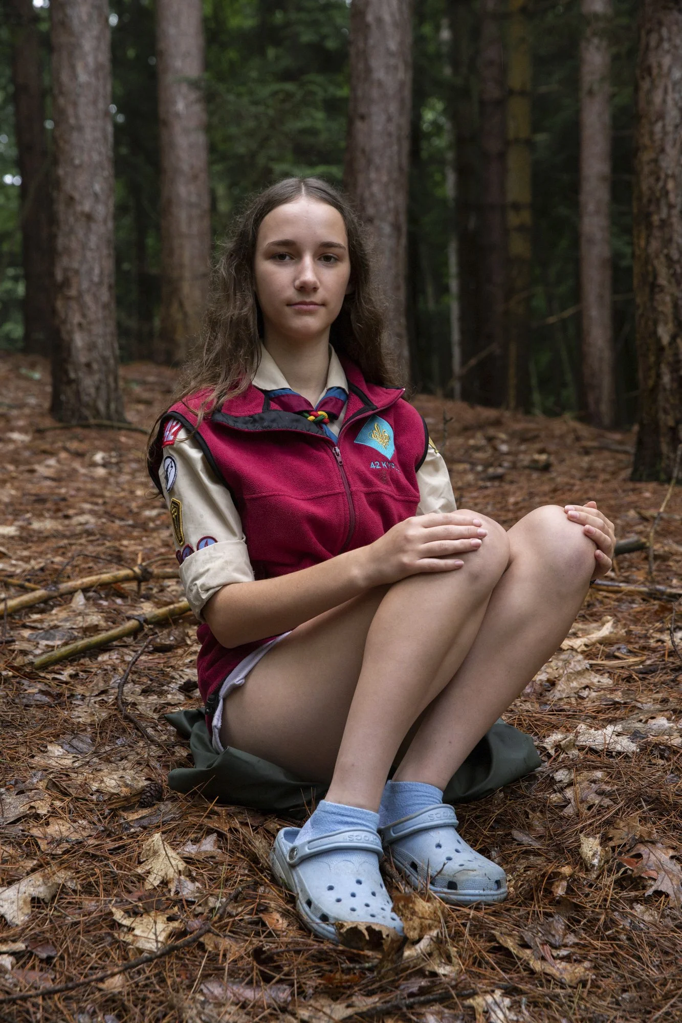 A serene young female Caucasian scout sits crossed-legged on the forest floor, wearing her vest adorned with badges, against a backdrop of tall trees.