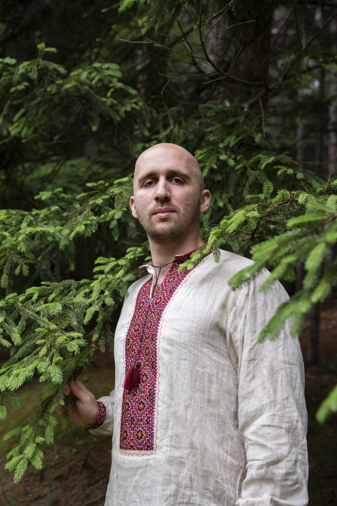 A young male Caucasian stands confidently in the woods looking at the camera, wearing a traditional Ukrainian embroidered shirt.
