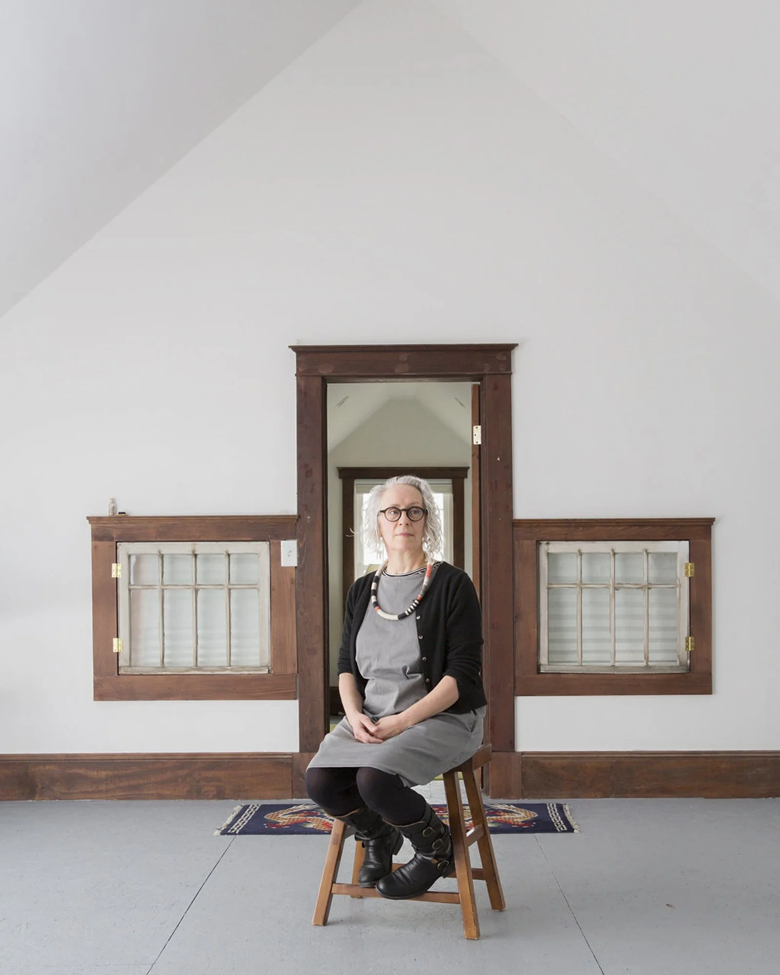 A Caucasian woman with glasses sits confidently in front of an ornate doorway, her serene posture matching the minimalist aesthetic of the room.
