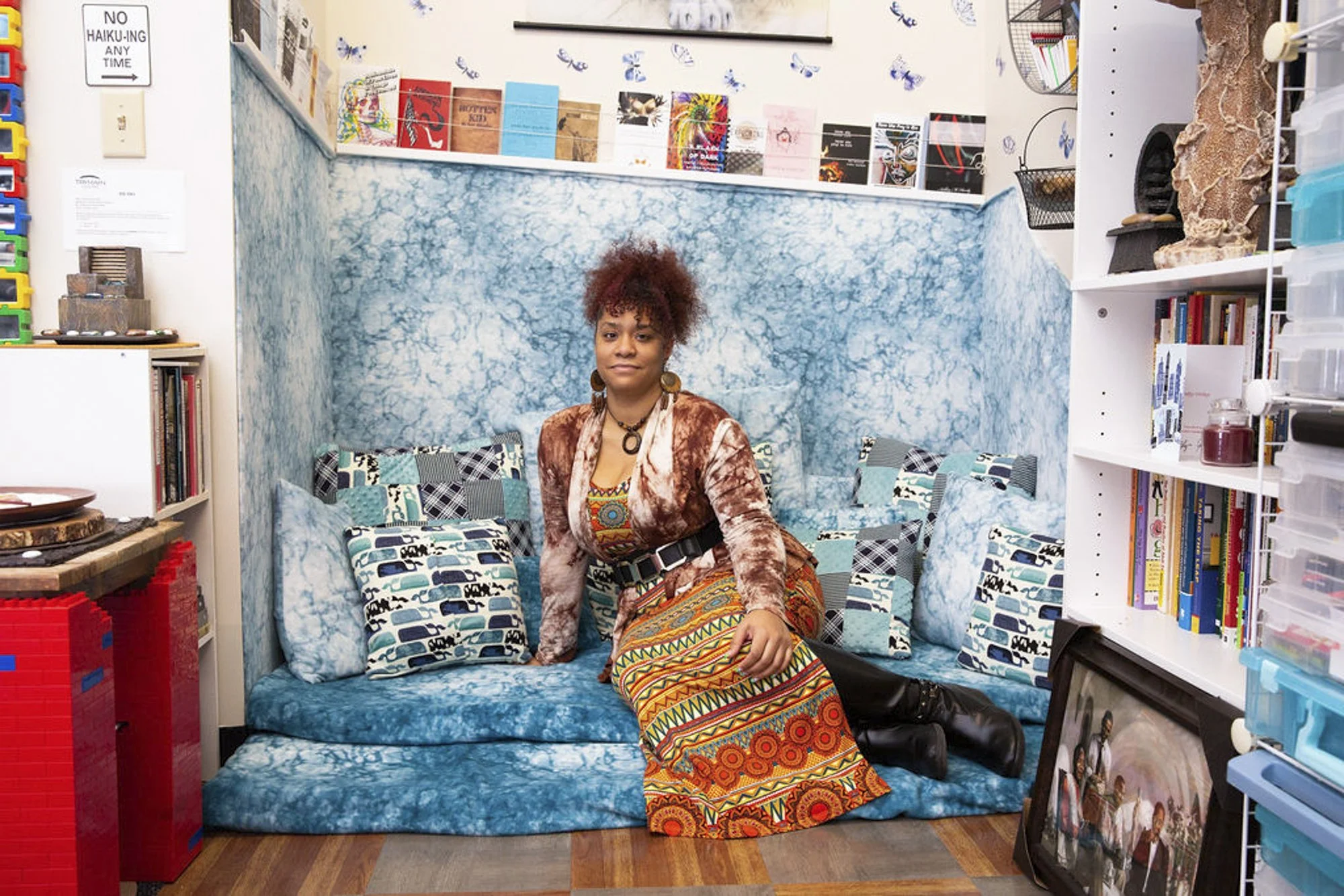 A young African American woman with a radiant smile sits on a blue couch, her unique personal style complementing the vibrant, eclectic decor around her.