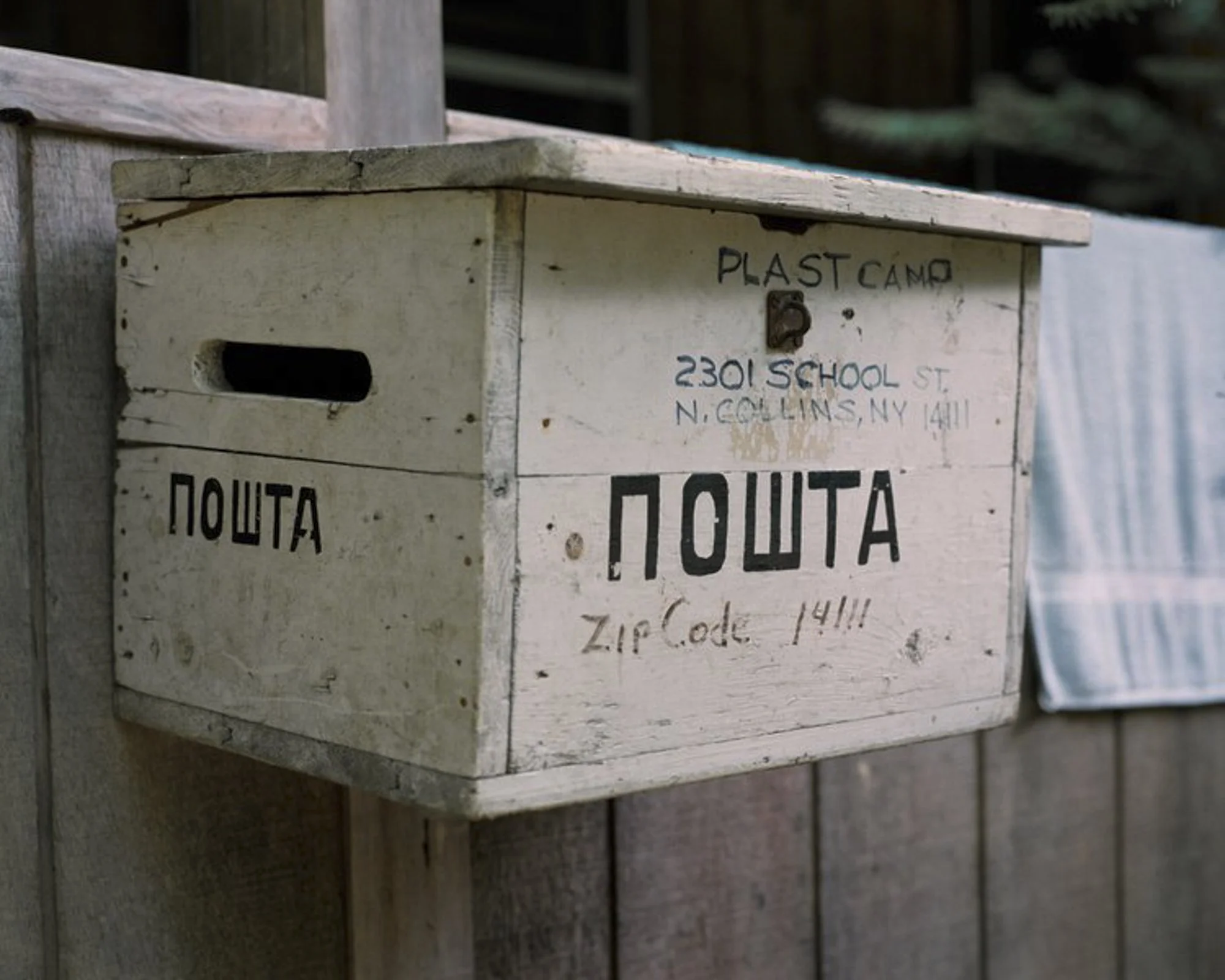 This is an image of a wooden mailbox with Cyrillic writing that reads "ПОШТА" (meaning "mail" in Ukrainian) and a U.S. address written above it. The text refers to "PLAST CAMP" and a location in North Collins, NY, indicating it might be part of a Uk
