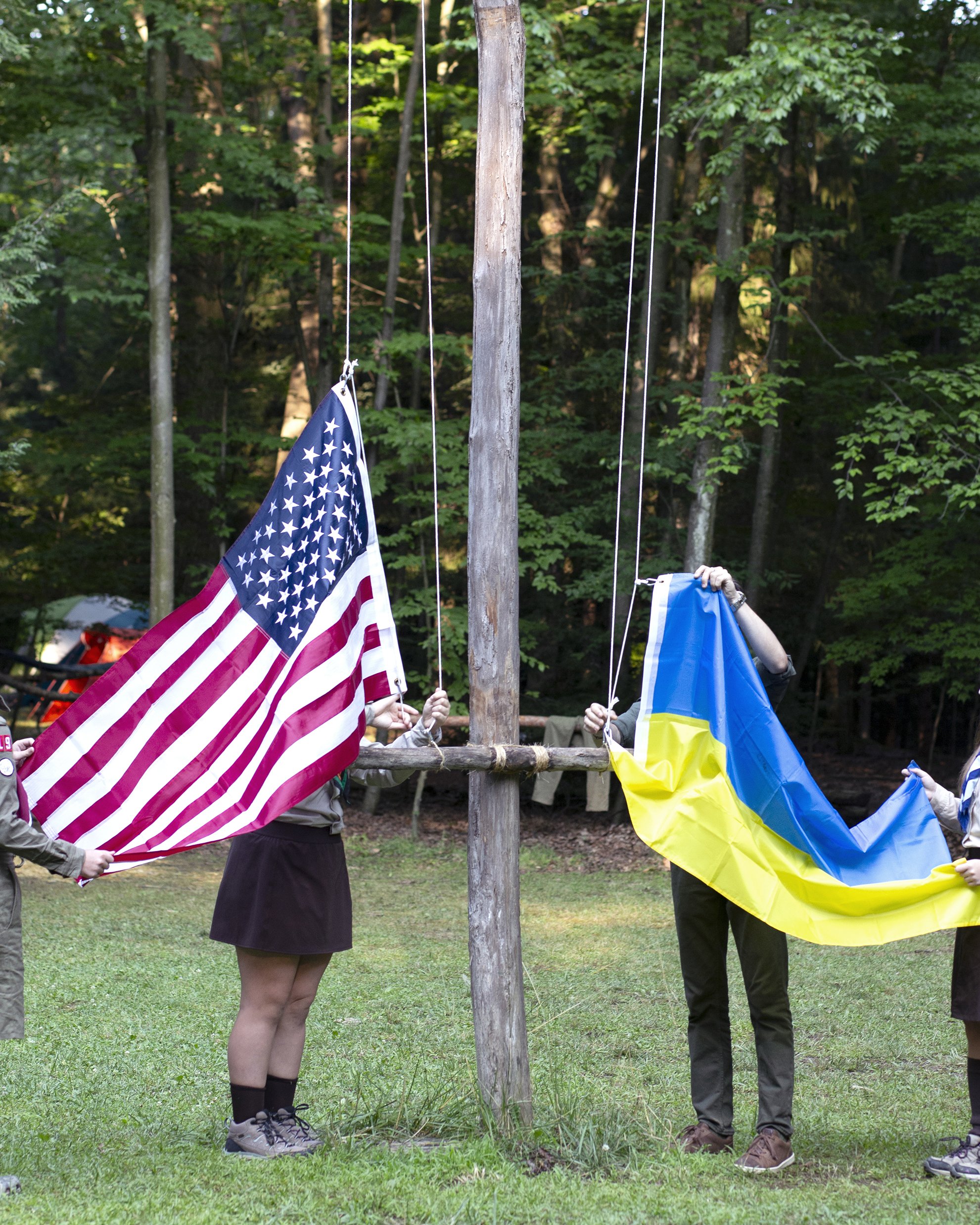 Two scouts in uniform raise an American flag and a Ukrainian flag simultaneously. Their faces are obscured by the flag.
