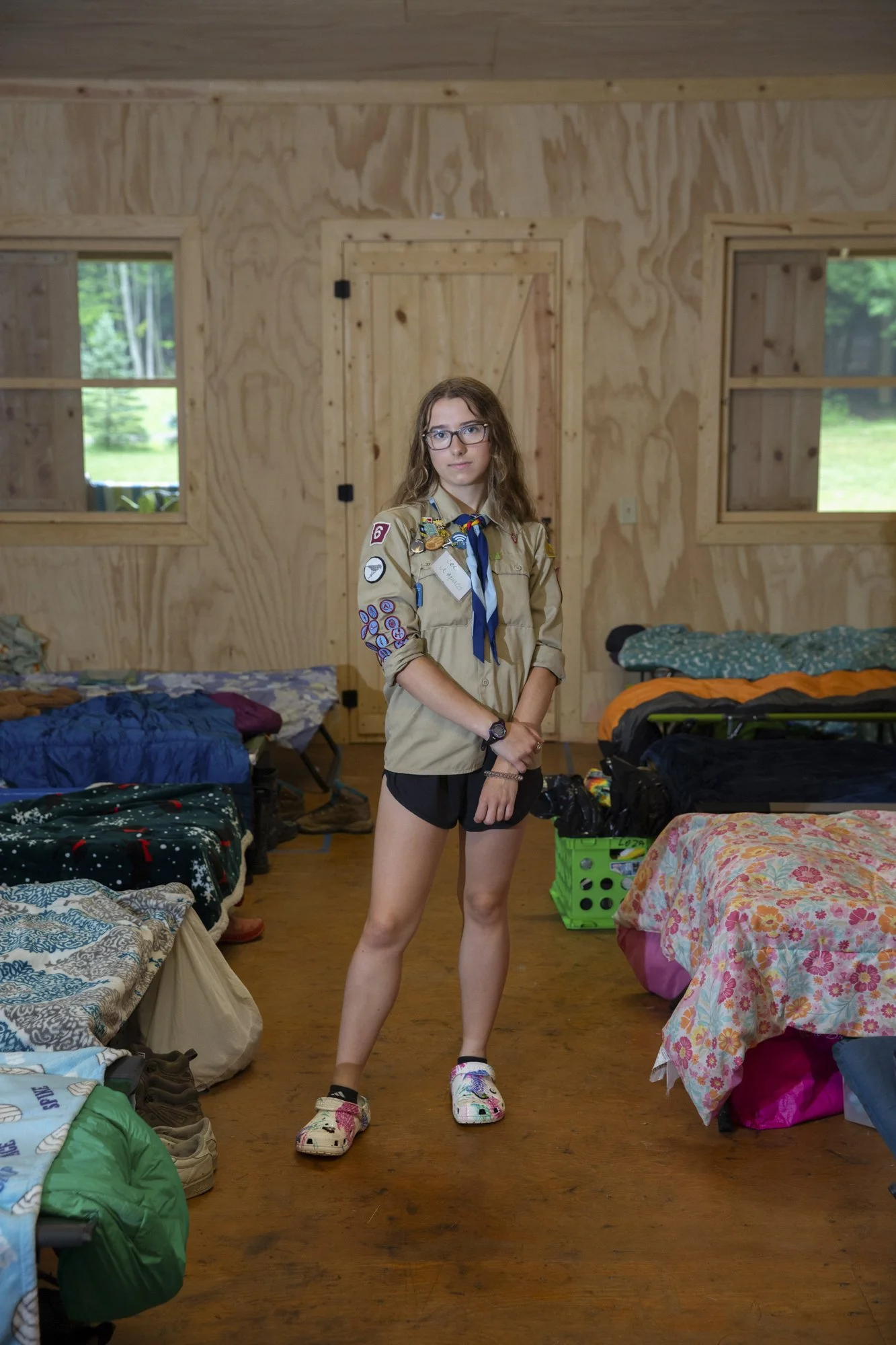 A young female Caucasian scout stands confidently in a cabin, surrounded by cots and sleeping bags.