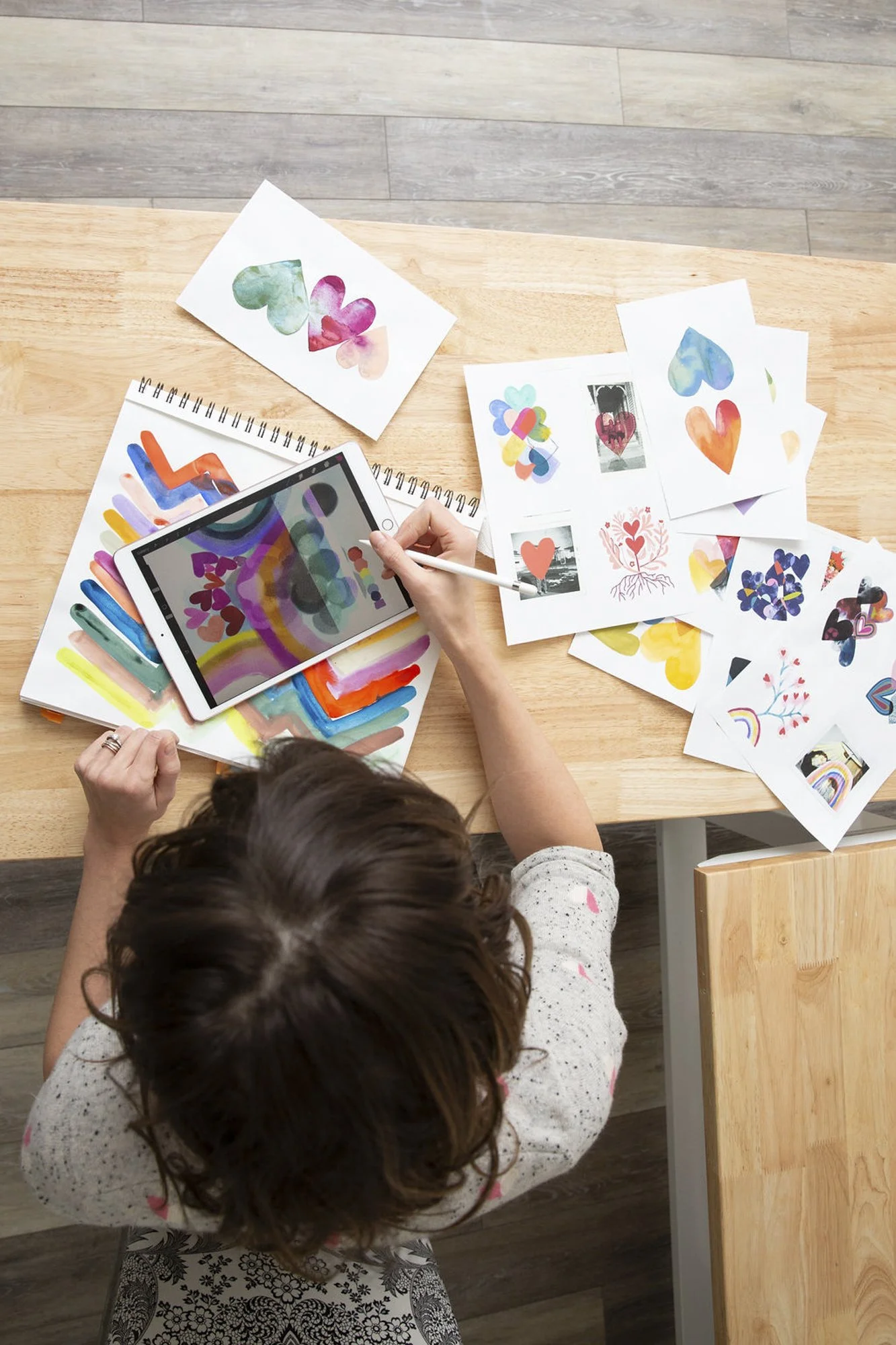 A top-down view of a young white girl creating colorful art on a digital tablet amidst a collection of vibrant drawings scattered on a wooden desk.