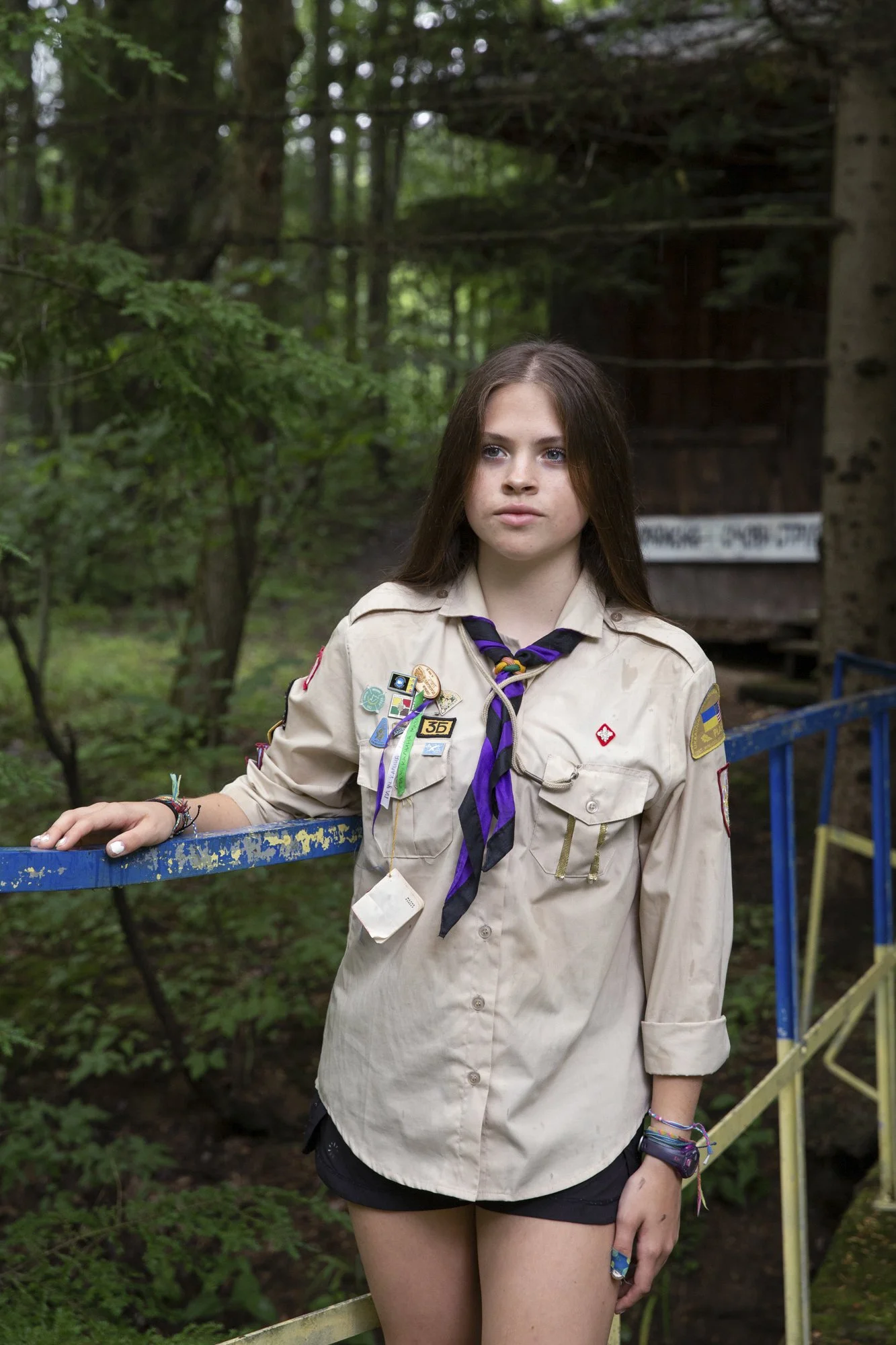 A young Caucasian female scout in uniform stands confidently in the woods, with badges displayed on her shirt.