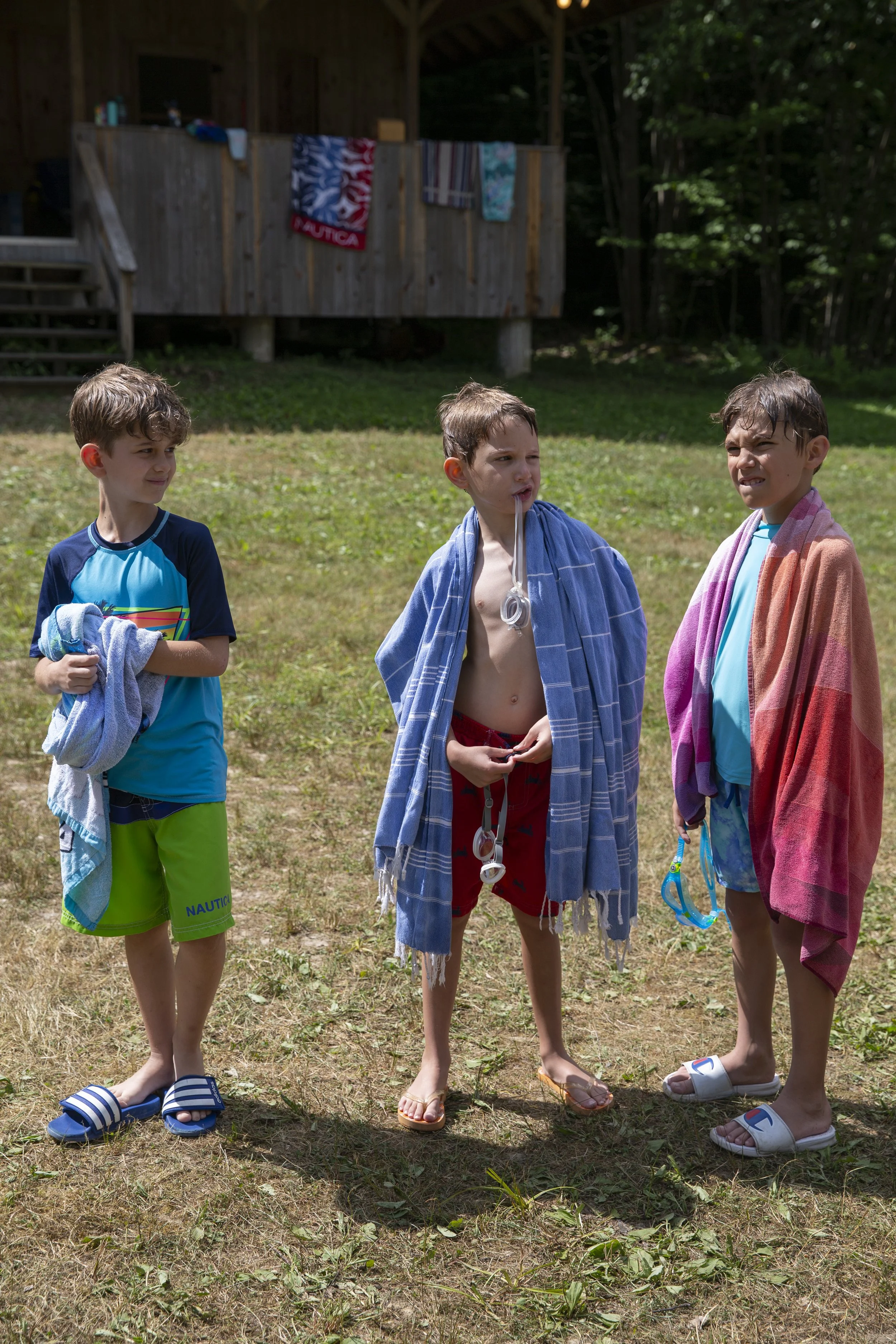 
ChatGPT
The image shows three young boys, approximately 6-8 years old, standing outside after what appears to be a swimming session. They are wearing swim trunks and towels over their shoulders, with goggles in hand or around their necks, suggesting