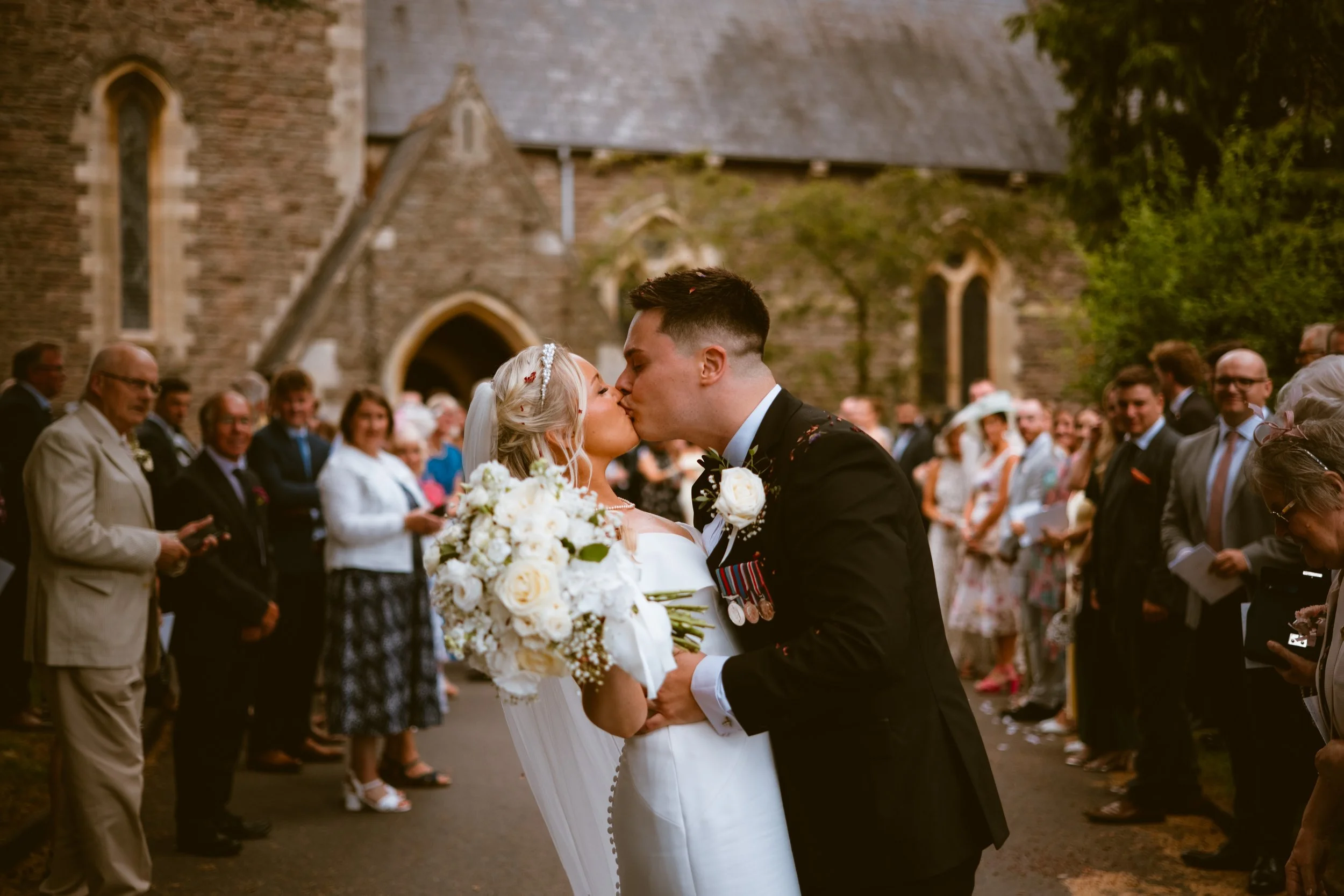 Newly married couple kissing outside a Herefordshire church surrounded by their guests