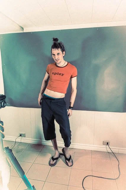 Young woman with dark hair in a high bun, wearing a red t-shirt with 'spicy' printed on it, dark pants, flip-flops, standing in front of a green chalkboard in a classroom or studio setting.