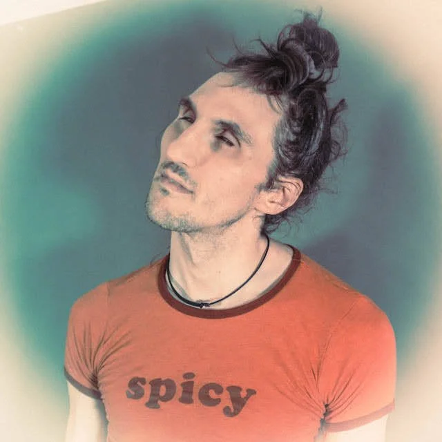 A young man with dark, curly hair styled in a bun, wearing a red t-shirt with the word "spicy" printed on it, and a black necklace, looking slightly to the side with a contemplative expression.