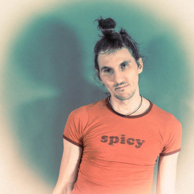 Young person with long hair tied in a top knot, wearing a red T-shirt with the word 'spicy' printed on it, standing against a green background.