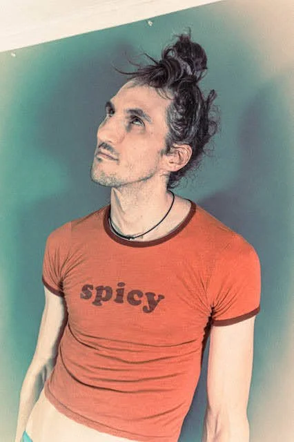 Man with long, messy hair tied in a bun, wearing an orange T-shirt with 'spicy' written on it, standing against a blue-green background.