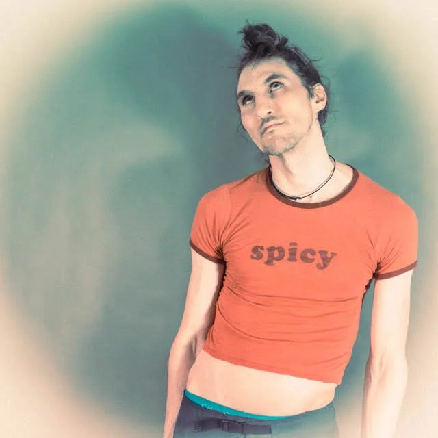 Person with dark hair tied up in a bun, wearing a red crop top with the word "spicy" on it, standing against a greenish background.