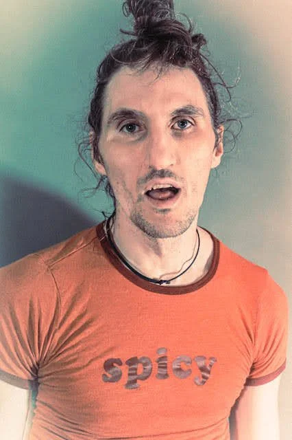 A man with long, curly hair tied up in a bun, wearing an orange T-shirt with the word 'spicy' printed on it, standing against a multicolored background.