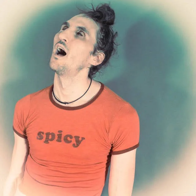 A man with dark, messy hair styled in a bun, wearing a red T-shirt with 'spicy' written on it, and a black necklace, posing with his head tilted back and mouth open.