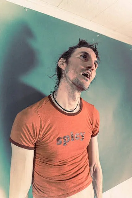 A man with long, dark, messy hair and a beard, wearing a red t-shirt with the word 'spice' on it, and a black necklace, standing against a teal wall, looking upward with an expressive face.