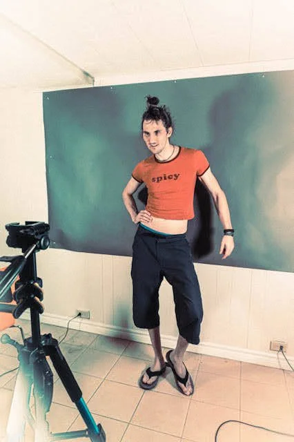 Young woman standing in front of a green screen, posing with one hand on her hip, wearing a red crop top, black pants, and flip-flops.