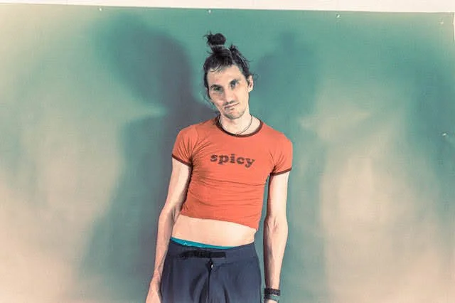 Young woman with dark hair in a bun, wearing a red crop top with 'spicy.' written on it, and dark pants, standing against a green wall.