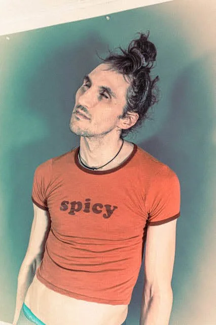 A person with long, messy hair styled in a top knot, wearing a red crop top with the word 'spicy' on it, and a black necklace, standing against a blue green background.