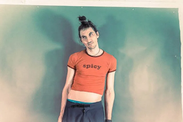 A young woman with dark hair tied in a messy bun, standing against a green wall. She is wearing a red crop top with the word "spicy" written on it, and black pants. She has a relaxed, slightly tilted head pose.